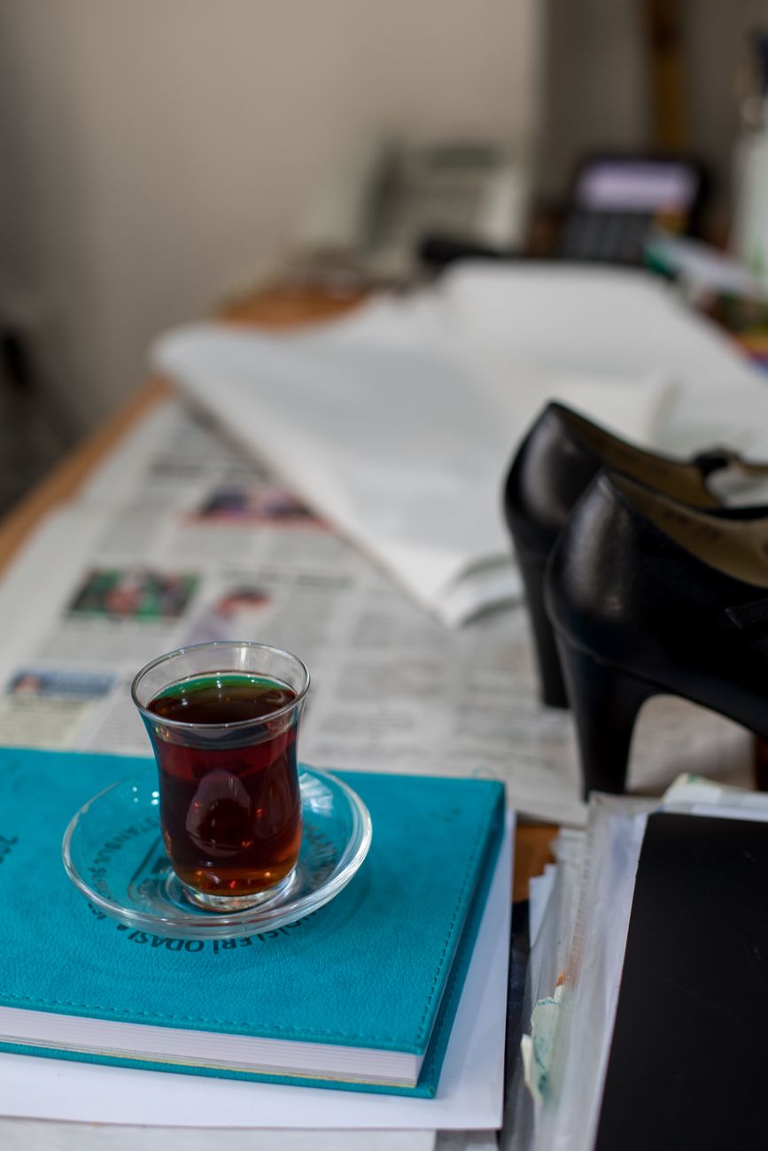 High angle of glass with drink with saucer on notebook and documents near heels and newspapers on table in light workspace